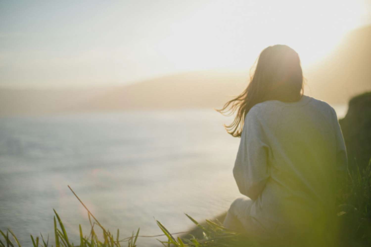 A woman with long, dark hair is sitting on a grassy cliff looking out at the ocean